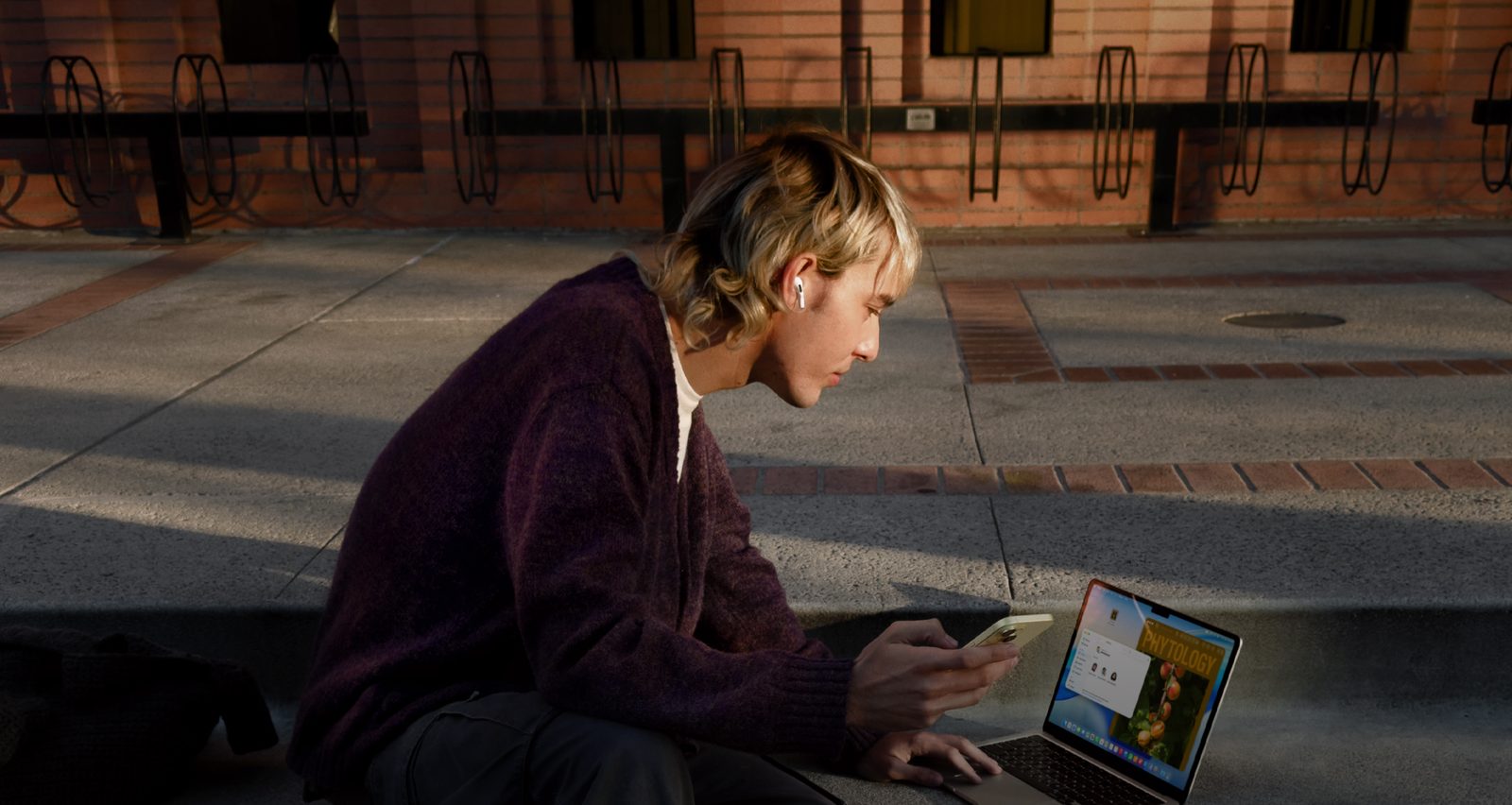 A student on campus steps working on a MacBook Air, AirPods in, iPhone in hand